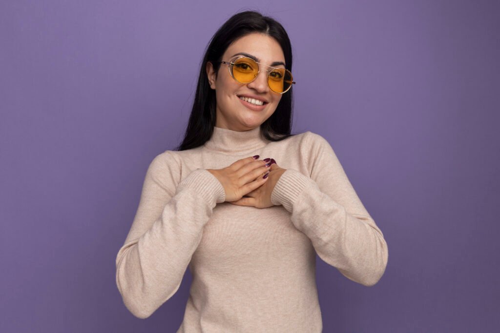 smiling pretty brunette caucasian girl in sun glasses puts hands on chest isolated on purple background with copy space